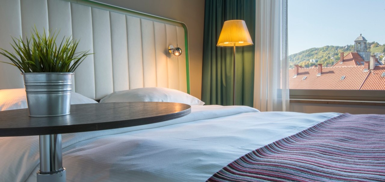 Modernes Hotelzimmer mit gemütlichem Bett, Nachttischlampe und Fensterblick auf rote Dächer und grüne Hügel. Ein Tisch mit Pflanze im Vordergrund., © Park Inn by Radisson Stuttgart