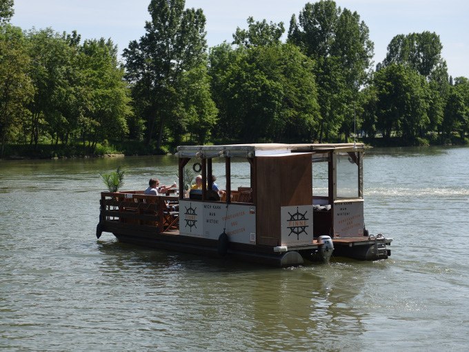 Ein rustikales Flo&szlig;boot mit &uuml;berdachtem Bereich f&auml;hrt auf einem Fluss. Im Hintergrund sind gr&uuml;ne B&auml;ume zu sehen., &copy; Cool-Tours StattReisen