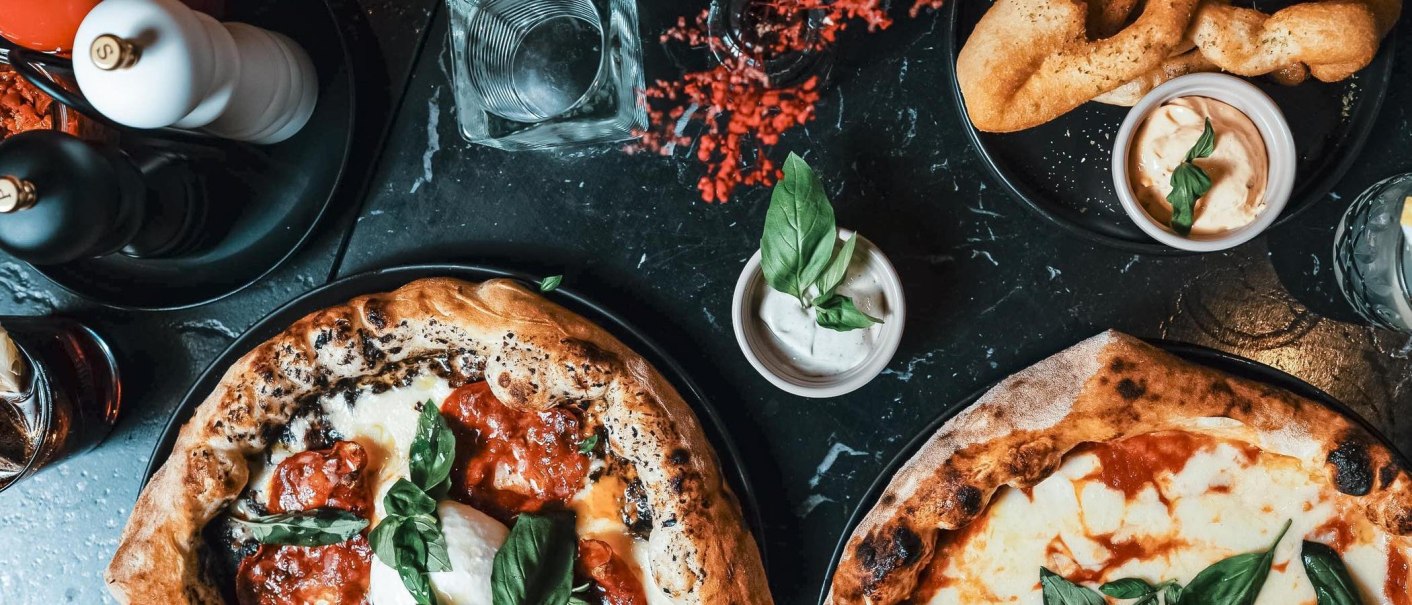 Von oben aufgenommenes Tablebild mit mehreren Pizzen, Dips, Gläsern und Gewürzmühlen auf dunklem Tisch., © Connective Media Von oben aufgenommenes Tablebild mit mehreren Pizzen, Dips, Gläsern und Gewürzmühlen auf dunklem Tisch., © Connective Media