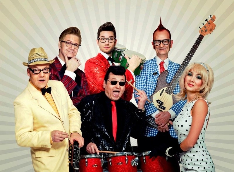 Sechs Personen in bunter, retro-inspirierter Kleidung posieren mit Musikinstrumenten vor einem strahlenförmigen Hintergrund., © C2 Concerts GmbH Sechs Personen in bunter, retro-inspirierter Kleidung posieren mit Musikinstrumenten vor einem strahlenförmigen Hintergrund., © C2 Concerts GmbH