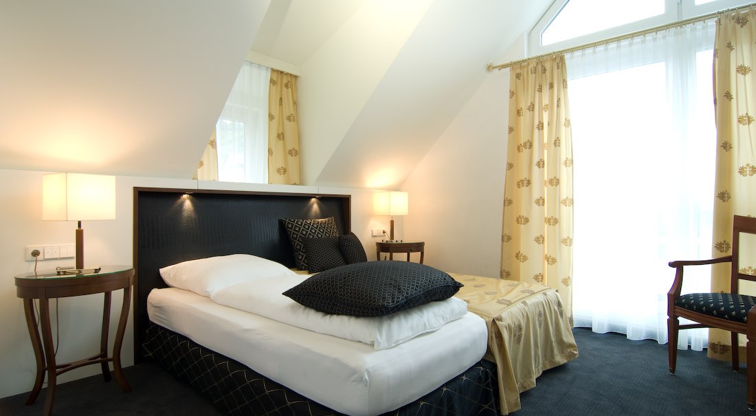 An elegant hotel room with a single bed, nightstands, and large windows with curtains. Light-colored walls and dark accents create a cozy atmosphere., © Jeanette Bender An elegant hotel room with a single bed, nightstands, and large windows with curtains. Light-colored walls and dark accents create a cozy atmosphere., © Jeanette Bender
