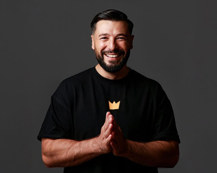 A man with a beard smiles at the camera. He wears a black T-shirt with a golden crown on it and holds his hands together., © Theaterhaus Stuttgart e.V. A man with a beard smiles at the camera. He wears a black T-shirt with a golden crown on it and holds his hands together., © Theaterhaus Stuttgart e.V.