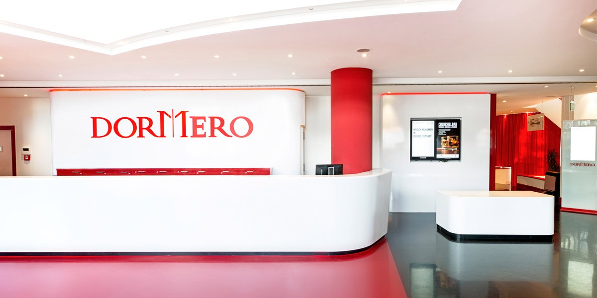 A modern hotel lobby with a white-and-red color scheme, the DORMERO logo on the wall, a screen displaying information, red carpet, and bright lighting., © Dormero Hotel Stuttgart A modern hotel lobby with a white-and-red color scheme, the DORMERO logo on the wall, a screen displaying information, red carpet, and bright lighting., © Dormero Hotel Stuttgart