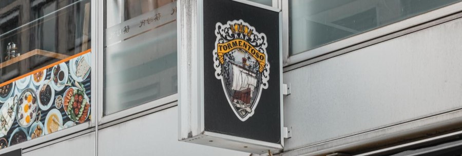 A sign with the Cape Tormentoso logo hangs on a modern building. It shows a sailing ship and decorative elements., © Stuttgart Marketing GmbH, Sarah Schmid