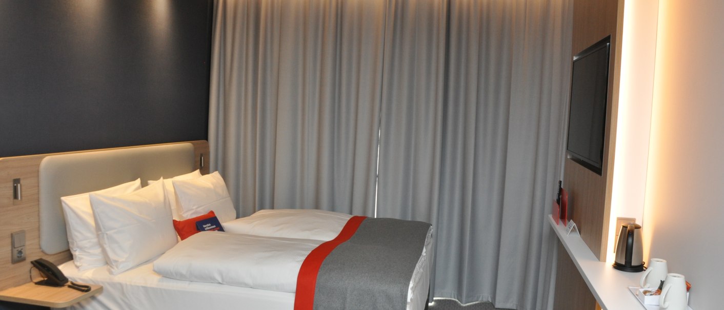 Modern hotel room with a double bed, gray curtains and a TV. A tray with cups and kettle stands on a shelf., © tristar GmbH Modern hotel room with a double bed, gray curtains and a TV. A tray with cups and kettle stands on a shelf., © tristar GmbH