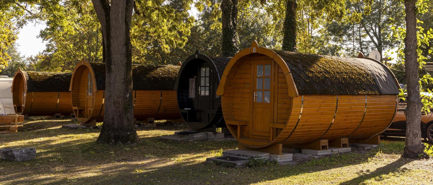 Fassförmige Holzhütten auf einem Campingplatz, umgeben von Bäumen und Sonnenlicht. Die Hütten stehen auf einem grasbewachsenen Boden., © Stuttgart-Marketing GmbH, Sarah Schmid Fassförmige Holzhütten auf einem Campingplatz, umgeben von Bäumen und Sonnenlicht. Die Hütten stehen auf einem grasbewachsenen Boden., © Stuttgart-Marketing GmbH, Sarah Schmid