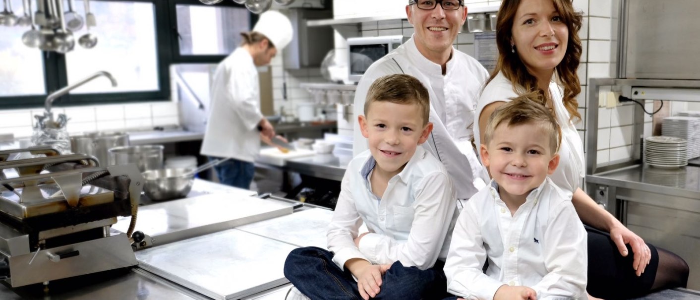 Familie in einer Restaurantküche, zwei Kinder sitzen auf einem Tisch, Eltern dahinter, Koch im Hintergrund., © Restaurant & Hotel Traube Hanweiler