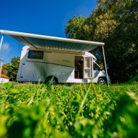 Ein Wohnmobil mit ausgefahrener Markise steht auf einer grünen Wiese unter blauem Himmel. Im Hintergrund sind Bäume und ein Gebäude zu sehen., © SMG, Thomas Niedermüller