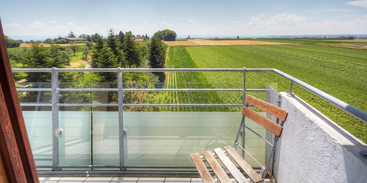 Ein Balkon mit Holzstuhl bietet einen weiten Blick auf grüne Felder und Bäume unter blauem Himmel., © Kemnater Hof GmbH