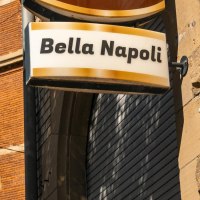 Ein Schild mit der Aufschrift 'Bella Napoli' h&auml;ngt an einem Backsteingeb&auml;ude. Der Himmel ist klar und blau, ein Baum ist im Vordergrund sichtbar., &copy; SMG, Sarah Schmid