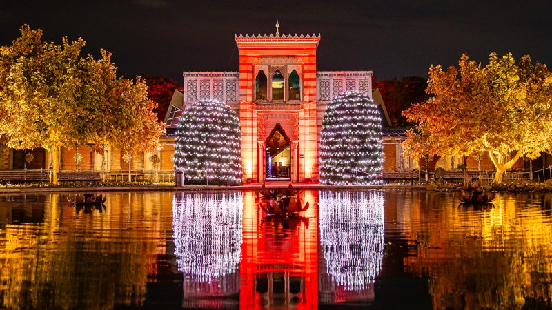 Ein beleuchtetes Gebäude mit roten und weißen Lichtern, umgeben von beleuchteten Bäumen, spiegelt sich in einem Teich bei Nacht., © Christmas Garden Rainer Keuenhof