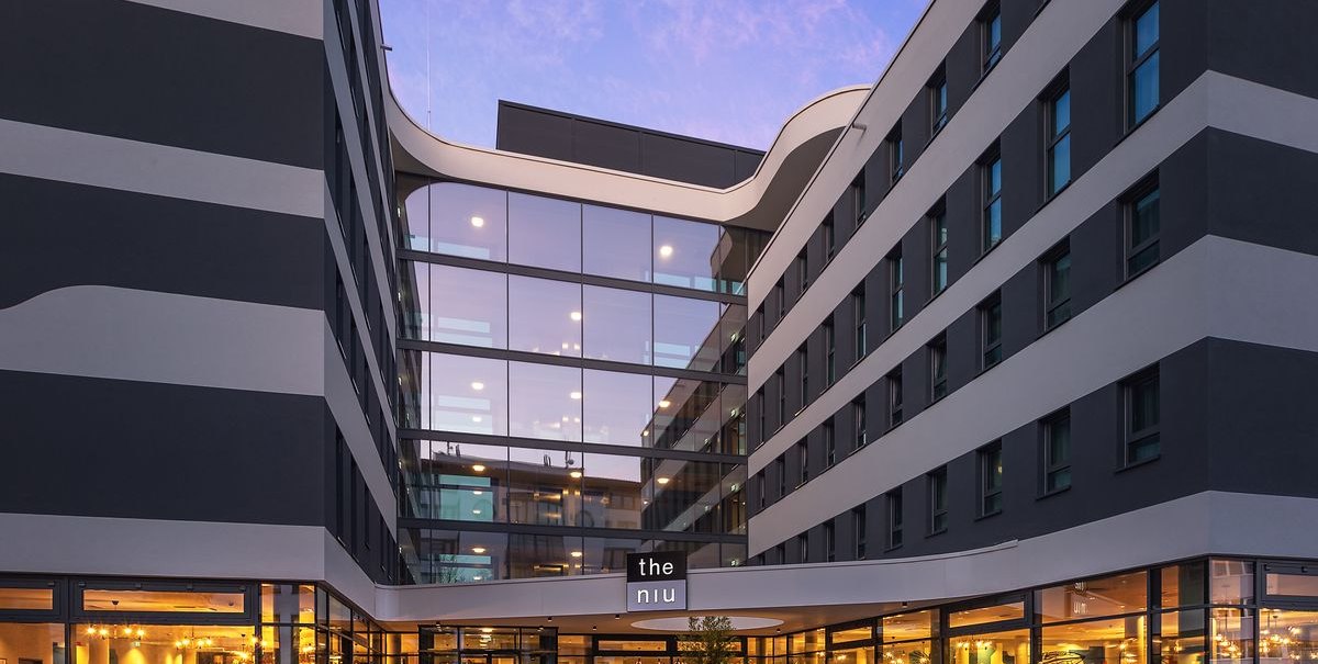 A modern hotel exterior with a glass facade and an illuminated entrance area at dusk. The building features an elegant, wave-like design., &copy; Novum Hospitality