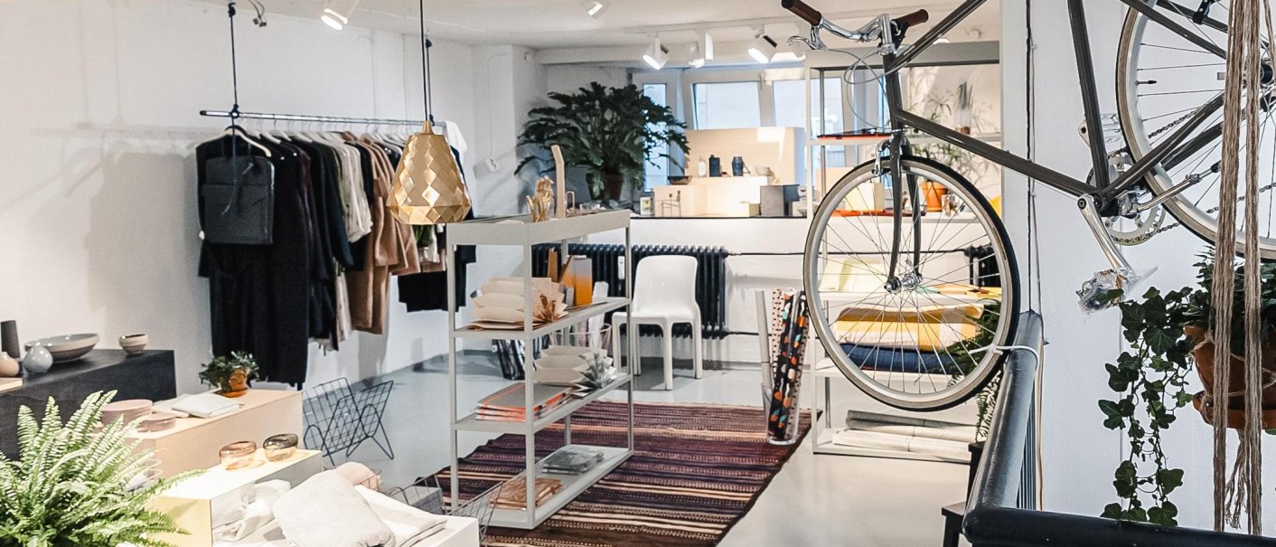 Moderner Laden mit Kleidung, Dekoration und einem Fahrrad an der Wand. Helle Beleuchtung und Pflanzen schaffen eine einladende Atmosphäre., © Kauf Dich Glücklich GmbH