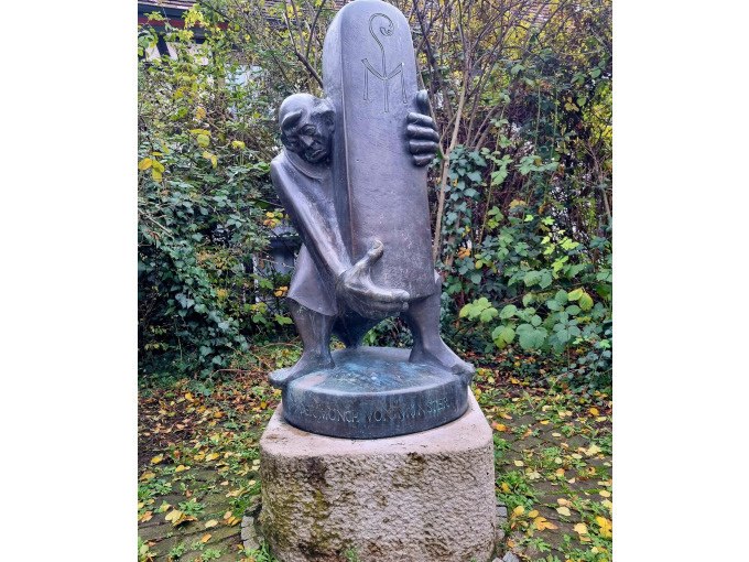 Bronzestatue eines Mannes, der eine große Steintafel umarmt, auf einem Sockel im Freien, umgeben von Laub und Büschen., © Cool-Tours StattReisen Bronzestatue eines Mannes, der eine große Steintafel umarmt, auf einem Sockel im Freien, umgeben von Laub und Büschen., © Cool-Tours StattReisen