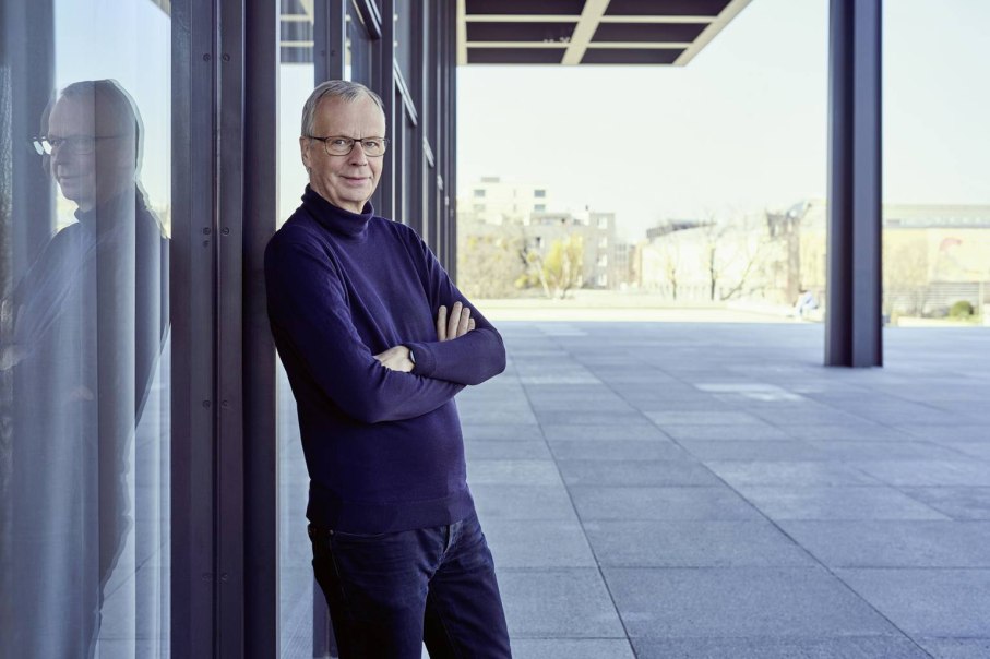 Ein Mann steht mit verschränkten Armen an einer Glaswand, seine Spiegelung ist sichtbar. Der Hintergrund zeigt eine moderne, urbane Umgebung., © Norman Posselt Ein Mann steht mit verschränkten Armen an einer Glaswand, seine Spiegelung ist sichtbar. Der Hintergrund zeigt eine moderne, urbane Umgebung., © Norman Posselt