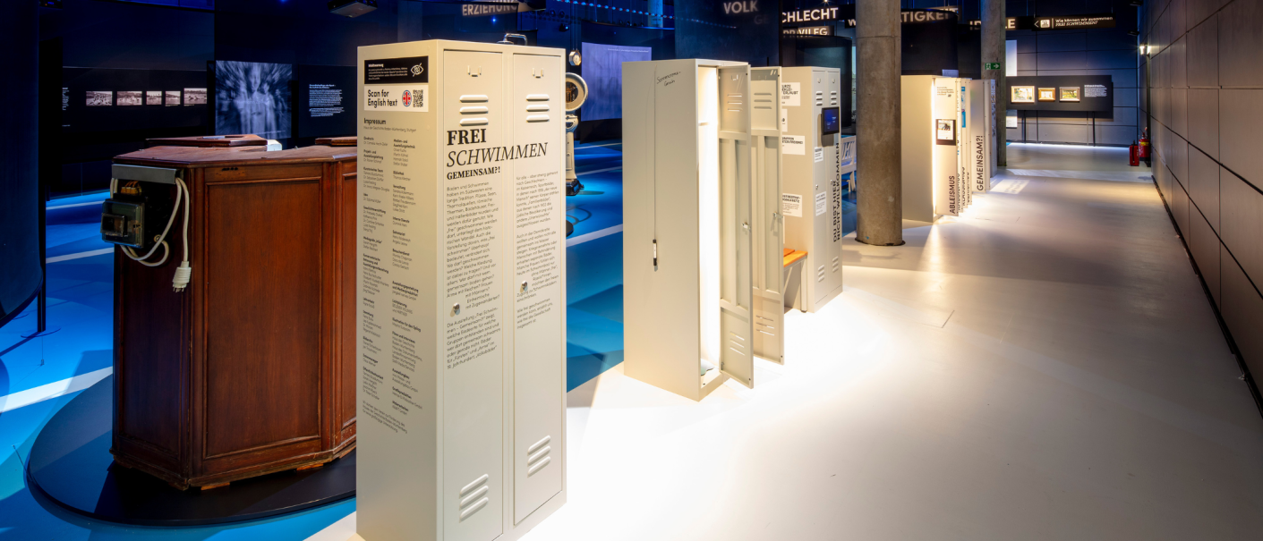 Museumsausstellung mit Schließfächern und Infotafeln zum Thema Schwimmen. Der Raum ist modern gestaltet und gut beleuchtet., © Frei_Schwimmen_Daniel_Stauch Museumsausstellung mit Schließfächern und Infotafeln zum Thema Schwimmen. Der Raum ist modern gestaltet und gut beleuchtet., © Frei_Schwimmen_Daniel_Stauch