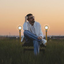 Eine Person sitzt nachdenklich auf einem Stuhl in einem Feld bei Sonnenuntergang, umgeben von zwei leuchtenden Lampen., &copy; links im Bild