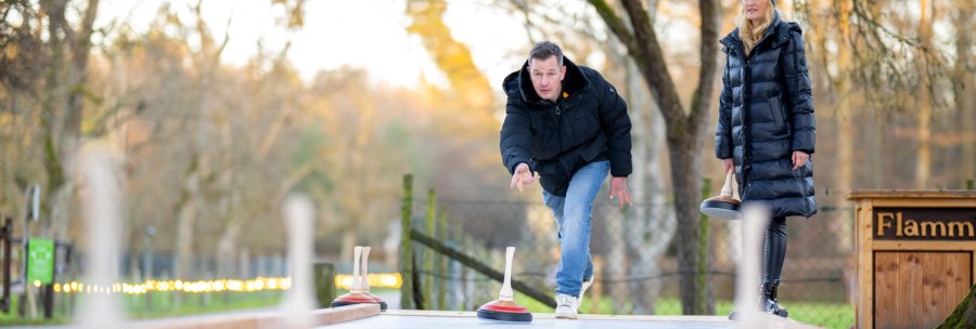 Ein Mann und eine Frau spielen Eisstockschie&szlig;en im Freien. Der Mann wirft gerade einen Eisstock, w&auml;hrend die Frau zuschaut. B&auml;ume im Hintergrund., &copy; Katzenbacher Hof