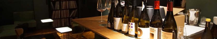 Several bottles of wine stand on a wooden table in a stylish bar. A record shelf and comfortable seating can be seen in the background., &copy; Weinhandlung Kreis KG