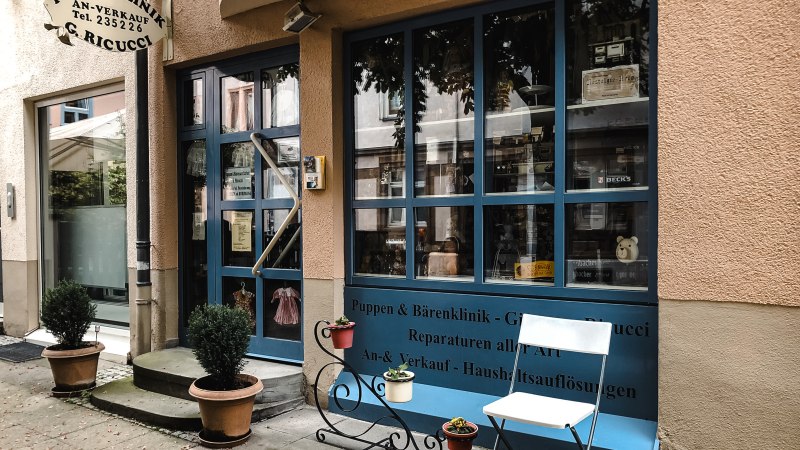 Eingang einer Puppenklinik mit blauer Tür und Schaufenster. Davor stehen Topfpflanzen und ein weißer Stuhl. Ein Schild weist auf Reparaturen hin., © Puppen- und Bärenklinik Eingang einer Puppenklinik mit blauer Tür und Schaufenster. Davor stehen Topfpflanzen und ein weißer Stuhl. Ein Schild weist auf Reparaturen hin., © Puppen- und Bärenklinik