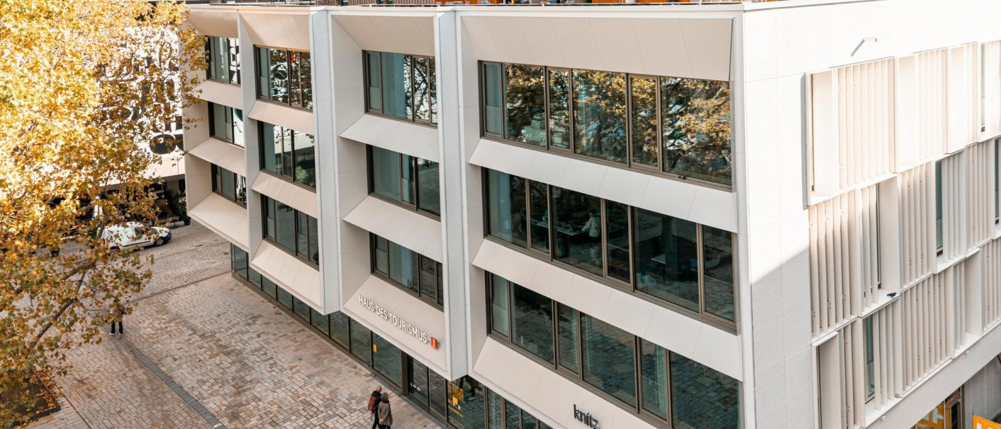 Modern façade of the House of Tourism with glass fronts and roof terrace. People stroll along the paved square in front of it., © SMG, Sarah Schmid Modern façade of the House of Tourism with glass fronts and roof terrace. People stroll along the paved square in front of it., © SMG, Sarah Schmid