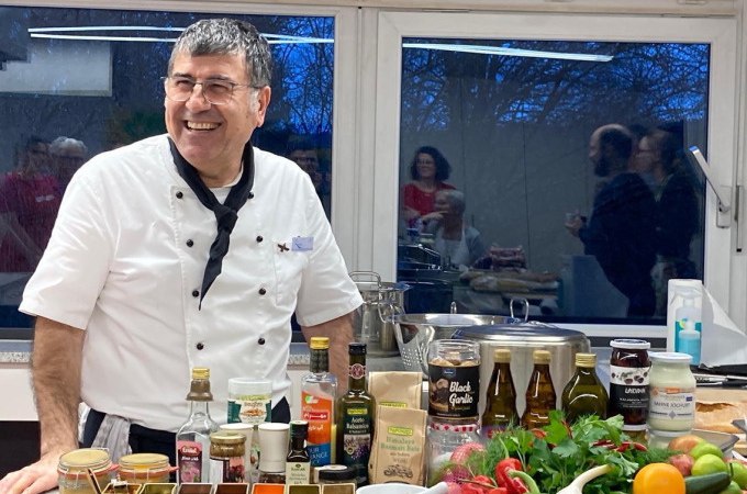 A chef in a white uniform presents a selection of ingredients and spices in a kitchen. People can be seen in the background., &copy; Kultweine-Kochwelten-Kunst&uuml;cke - Kochschule Sonja Lenz