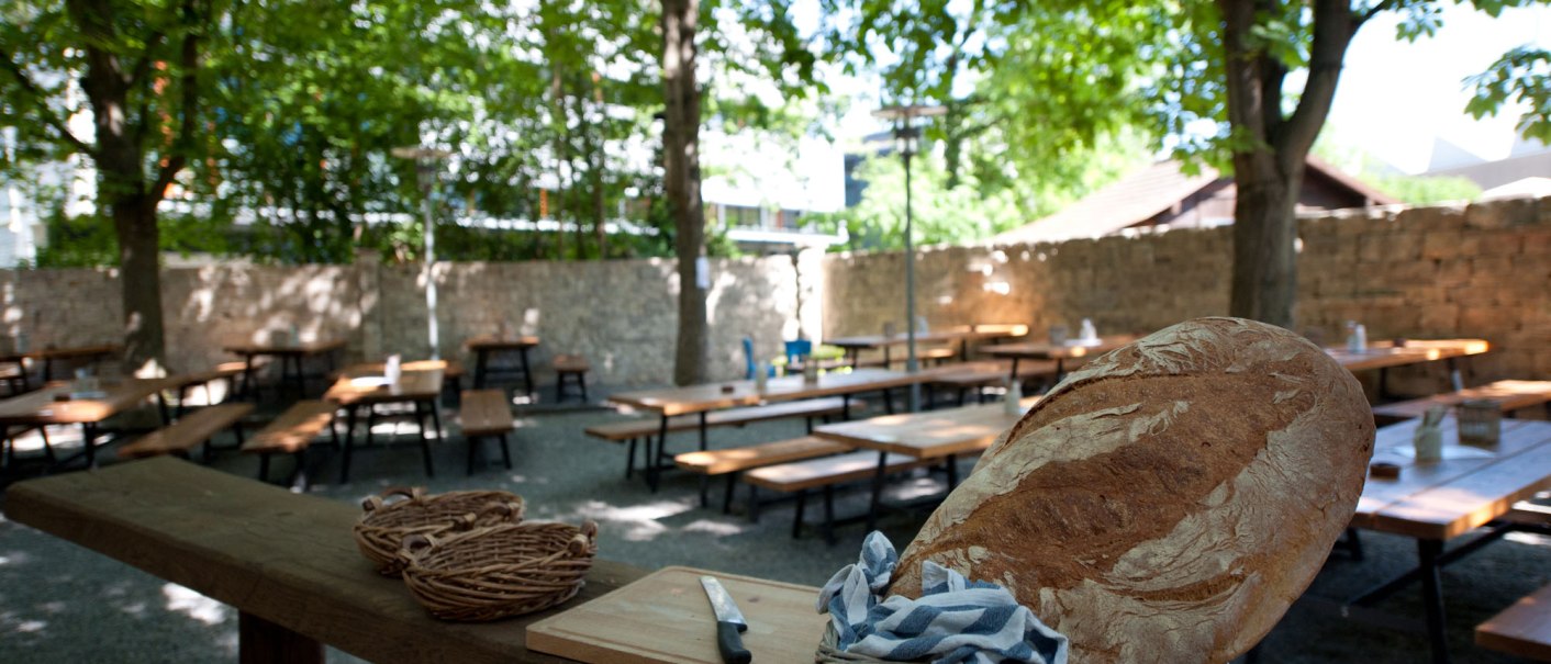 Ein leerer Biergarten mit Holztischen und Bänken unter Bäumen. Im Vordergrund ein Brotkorb mit einem Laib Brot und einem Messer., © Ruthenberg&Schlatter GbR Ein leerer Biergarten mit Holztischen und Bänken unter Bäumen. Im Vordergrund ein Brotkorb mit einem Laib Brot und einem Messer., © Ruthenberg&Schlatter GbR