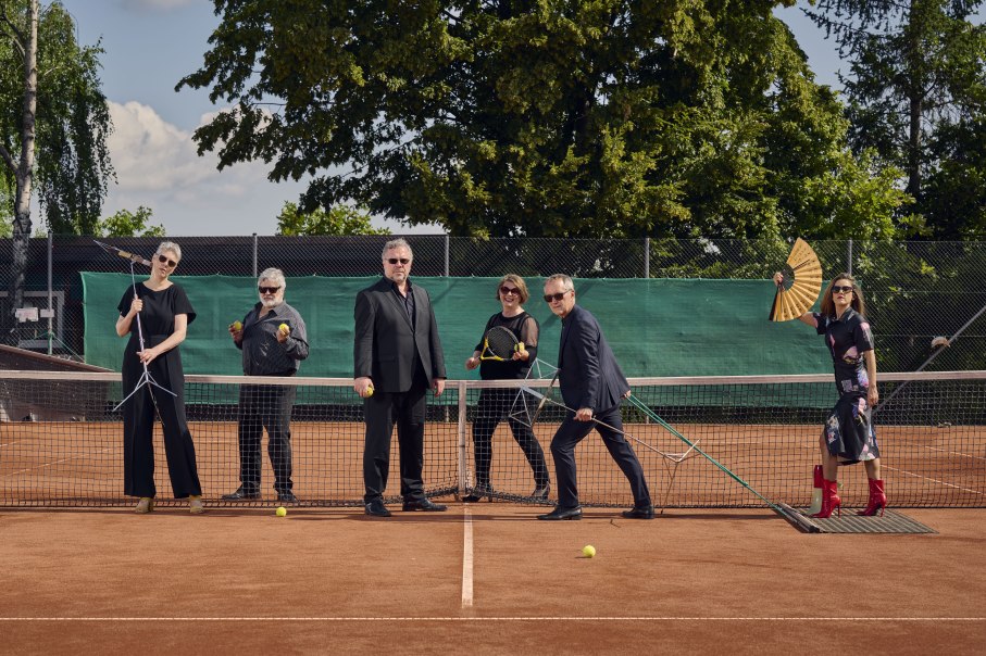 Sechs Personen posieren auf einem Tennisplatz mit Tennisschl&auml;gern und B&auml;llen. Eine Person h&auml;lt einen F&auml;cher. Sie stehen vor einem Netz, umgeben von B&auml;umen., &copy; Martin Sigmund