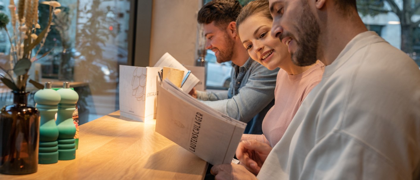 Three people are sitting at a table in a restaurant reading the menu. In the background is a window with a view outside., © Stuttgart-Marketing GmbH, Martina Denker