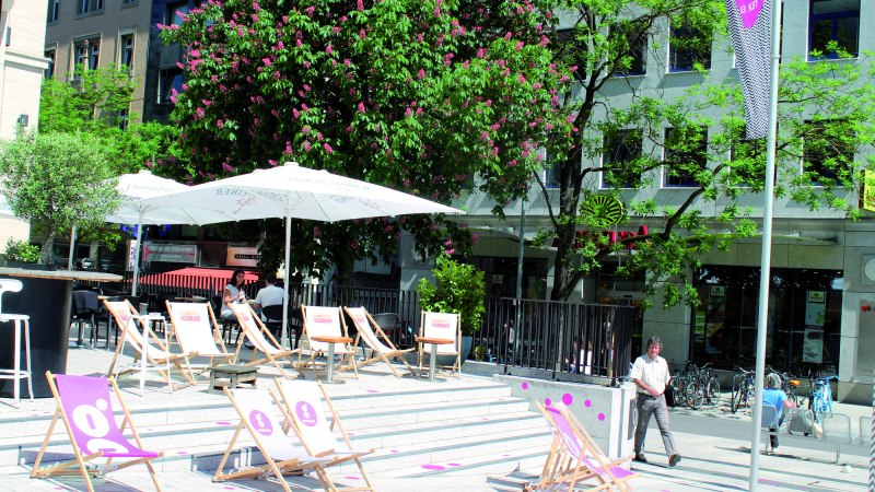 Sonniger Außenbereich mit Liegestühlen und Sonnenschirmen vor einem Gebäude. Blühende Bäume und Passanten im Hintergrund., © Stuttgart-Marketing GmbH (SMG)