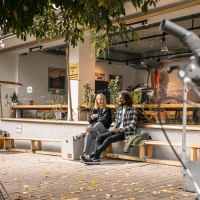 Zwei Personen sitzen lachend vor einem Café auf einer Bank. Im Vordergrund ist ein Fahrradlenker zu sehen. Herbstliche Blätter liegen auf dem Boden., © SMG Stuttgart Marketing GmbH - Sarah Schmid