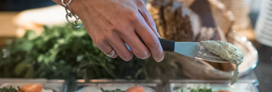 A hand with a wristband takes cream of mushroom from a bowl. Next to it are bowls of herb cream cheese and paprika cream, with bread in the background., © Café Justus, Stuttgart