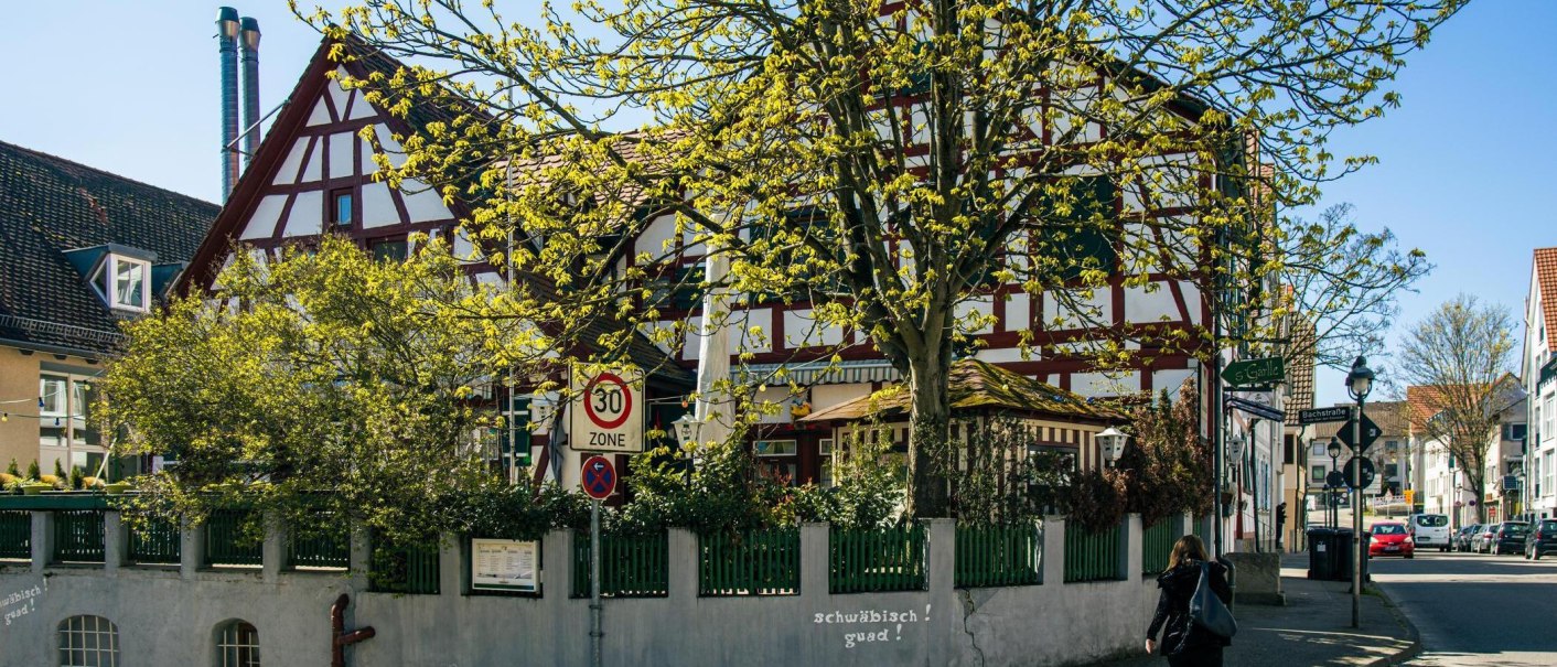 Half-timbered house in Kornwestheim, surrounded by trees and street signs. A person walks along the street while cars are parked., © Stuttgart-Marketing GmbH, Sarah Schmid