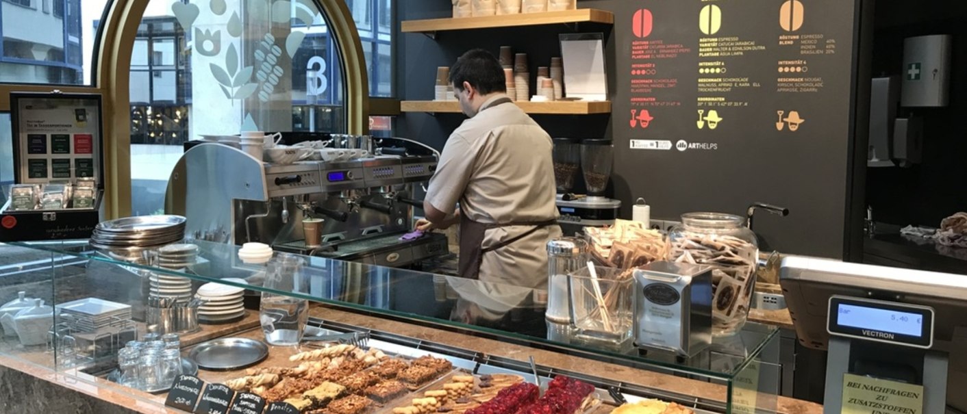 A barista in a caf&eacute; prepares coffee. In front of him are pastries, a coffee machine and packets of tea., &copy; Old Bridge, Stuttgart