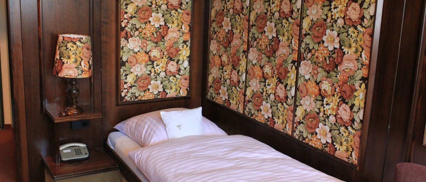 A hotel room with a single bed, floral wallpaper, and a matching lamp. A telephone sits on a small shelf., &copy; Hotel Spahr