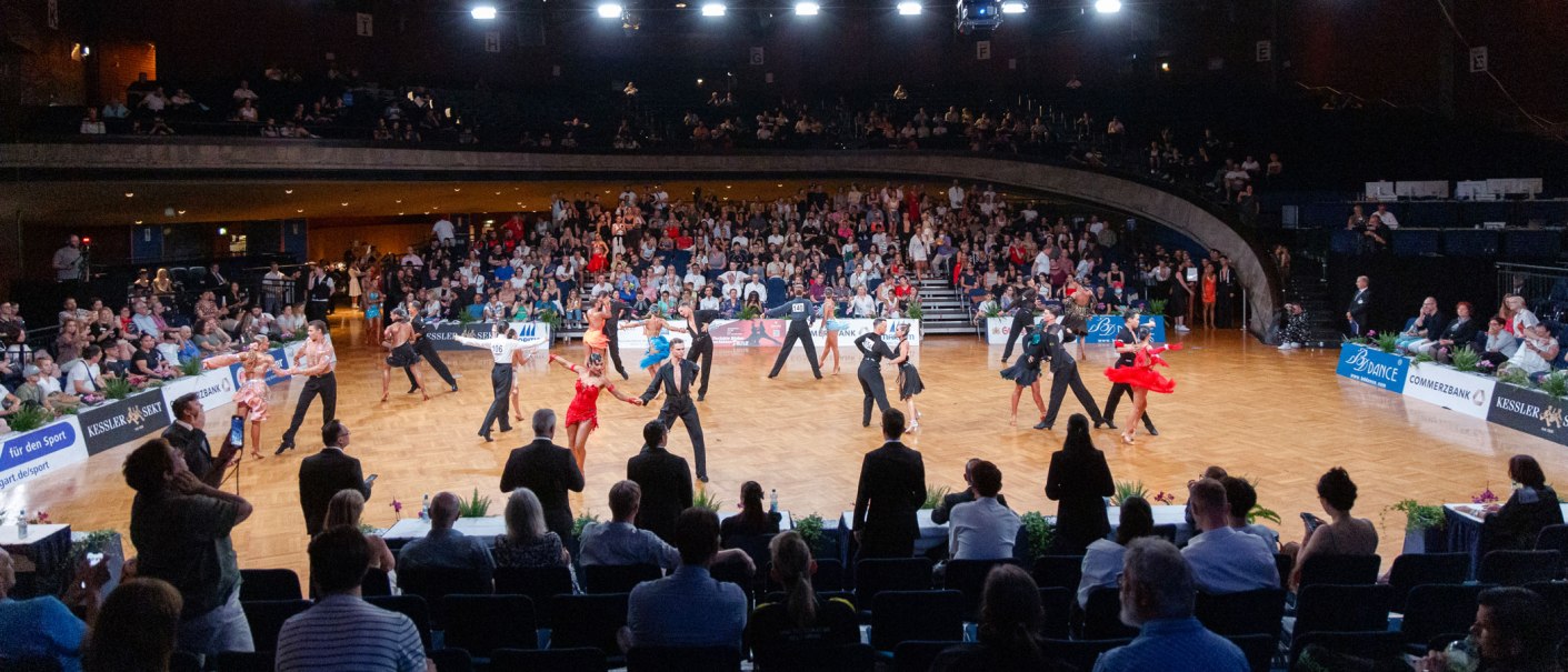 Dancing couples in elegant attire dance on a tournament floor, surrounded by a large audience in a hall., &copy; Bob van Ooik