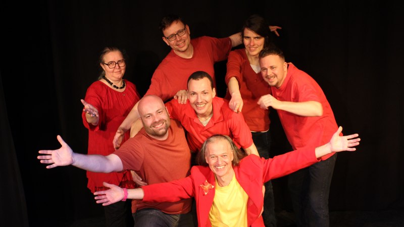Sieben Personen in roten Outfits posieren fröhlich auf einer Bühne. Sie lächeln und strecken die Arme aus. Hintergrund ist schwarz., © kaba-reh production Sieben Personen in roten Outfits posieren fröhlich auf einer Bühne. Sie lächeln und strecken die Arme aus. Hintergrund ist schwarz., © kaba-reh production