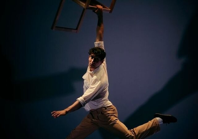 Ein Mann in einem wei&szlig;en Hemd und beigen Hosen h&auml;ngt an einem Stuhl in der Luft, umgeben von einem dunklen Hintergrund., &copy; Theaterhaus Stuttgart e.V.