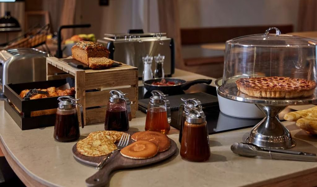 Ein Frühstücksbuffet mit verschiedenen Gebäcksorten, Sirupflaschen, Brot und einem Kuchen unter einer Glasglocke auf einem Tisch., © Adagio