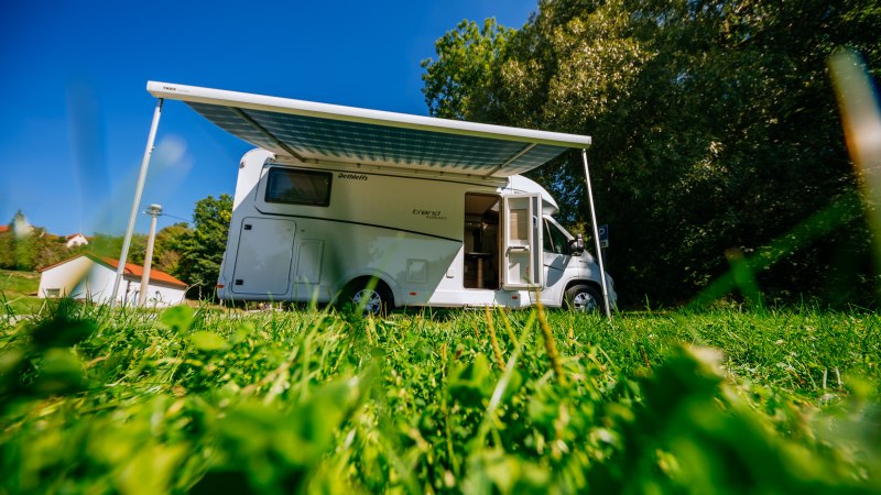 Ein Wohnmobil mit ausgefahrener Markise steht auf einer grünen Wiese unter blauem Himmel. Im Hintergrund sind Bäume und ein Gebäude zu sehen., © SMG, Thomas Niedermüller