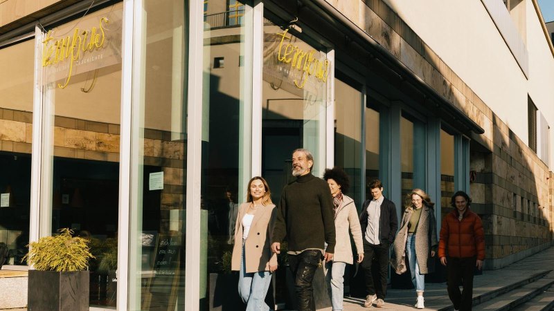 Eine Gruppe von Menschen verlässt das Tempus Café & Bistro, ein modernes Gebäude mit Glasfassade. Im Vordergrund stehen gestapelte Stühle und ein geschlossener Sonnenschirm., © Stuttgart-Marketing GmbH, Alwin Maigler