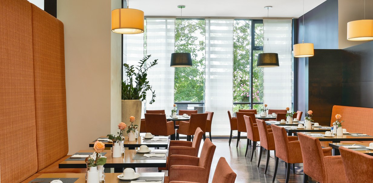 Modernes Restaurant mit orangefarbenen Stühlen, Tischen und Blumen. Große Fenster sorgen für viel Licht., © NH Hotel Stuttgart Airport