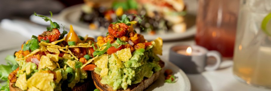Avocado toast with tomatoes and herbs, a sandwich and drinks in the background. A tea light creates a cozy atmosphere., © Veganes Gold®
