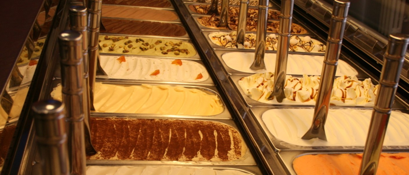 An ice cream counter with various flavors in metal containers, including chocolate, pistachio and vanilla. Metal spoons are placed in the containers., © Old Bridge, Stuttgart