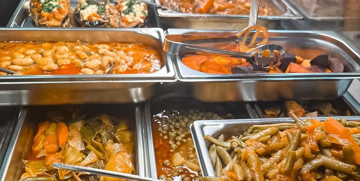 Ein Buffet mit verschiedenen warmen Speisen in Edelstahlbehältern, darunter Bohnen, Gemüse und gefüllte Auberginen., © Zauberküche, Stuttgart