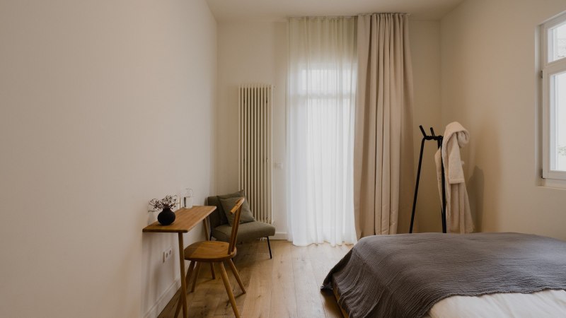 Minimalistisch eingerichtetes Schlafzimmer mit Holzboden, Bett, kleinem Schreibtisch und Stuhl. Vorhänge und ein Bademantel auf einem Ständer ergänzen das Bild., © Peter Oliver Wolff / DAS EDITH Minimalistisch eingerichtetes Schlafzimmer mit Holzboden, Bett, kleinem Schreibtisch und Stuhl. Vorhänge und ein Bademantel auf einem Ständer ergänzen das Bild., © Peter Oliver Wolff / DAS EDITH