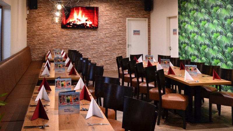 Ein langer Tisch in einer Lounge, dekoriert mit roten und wei&szlig;en Servietten. An der Wand ein Bildschirm mit Kaminfeuer, daneben eine gr&uuml;ne Tapete., &copy; Bowling & Billard Lounge
