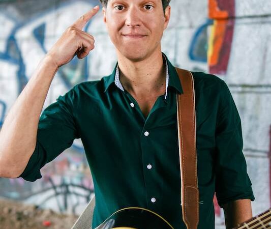 A man with a guitar stands in front of a graffiti wall and points to his head with his finger. He is wearing a dark green shirt and smiling slightly., &copy; Rosenau Kultur e.V.