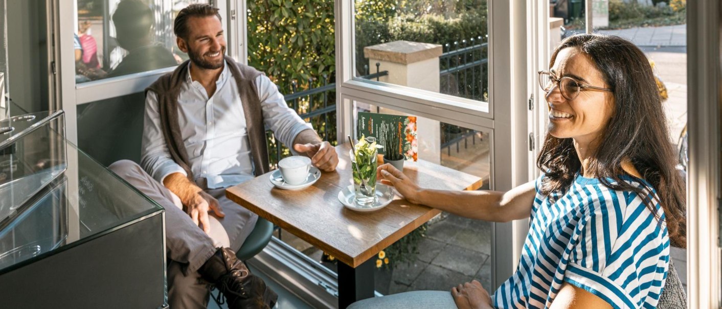 Zwei Personen sitzen in einem Caf&eacute; am Fenster, trinken Kaffee und unterhalten sich l&auml;chelnd. Sonnenlicht f&auml;llt durch das Fenster., &copy; Stuttgart-Marketing GmbH, Sarah Schmid