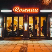 Eingang eines Lokals namens Rosenau mit leuchtendem Schild. Menschen sitzen drinnen, Plakate an den Wänden, gemütliche Atmosphäre., © Rosenau e. V., Stuttgart